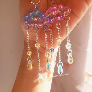 May include: Iridescent cloud-shaped earrings with dangling chains, hearts, stars, and teardrop-shaped crystals. The earrings have silver-colored chains and hooks. The earrings are a whimsical accessory.