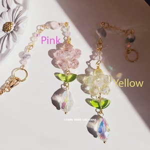 May include: Two floral-themed earrings with gold-colored chains. One earring is pink, and the other is yellow. Each earring features a flower, green leaves, and a crystal teardrop. The text "FAIRY TROVE CREATIONS" is visible.