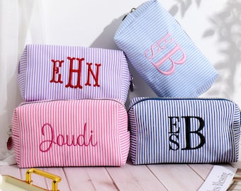 May include: Four striped fabric cosmetic bags with different embroidered monograms. The bags are in pink, purple, blue and navy blue.
