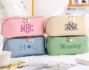 May include: Four different cosmetic bags stacked on top of each other. The top bag is pink with the monogram "MHC" embroidered on the front. The second bag is beige with the monogram "FSR" embroidered on the front. The third bag is blue with the monogram "HOG" embroidered on the front. The bottom bag is green with the word "Wesley" embroidered on the front.