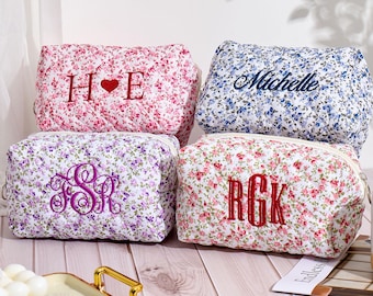May include: Four floral print cosmetic bags with different embroidered initials and names. The bags are arranged in a row on a wooden surface. The bags are white with a floral pattern in shades of pink, purple, and blue.