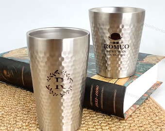 May include: Two stainless steel tumblers with a hammered texture. One tumbler has a monogram with the letters "D" and "R" inside a wreath. The other tumbler has the text "ROMEO BEST MAN 18-10-2023" with a hat graphic.