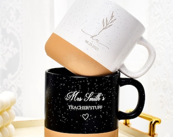 May include: Two ceramic mugs, one white and one black, stacked on a tray. The white mug has a tan base and a sprig design with text. The black mug has a tan base and the text "Mrs Smith's TEACHER'STUFF".