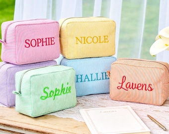 May include: A collection of personalized cosmetic bags in various colors, including pink, yellow, blue, green, and orange, each embroidered with a different name. The bags are striped and rectangular, with a zipper closure. A vase of white calla lilies and a notebook are also present.