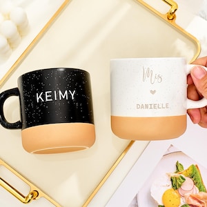 以下が含まれることがあります： 水玉模様のデザインのセラミックマグカップ2つ。1つは黒で、白で「KEIMY」と書かれており、もう1つは白で、「DANIELLE」という名前と筆記体で「Mis」と書かれています。どちらのマグカップもタン色のベースとハンドルが付いています。