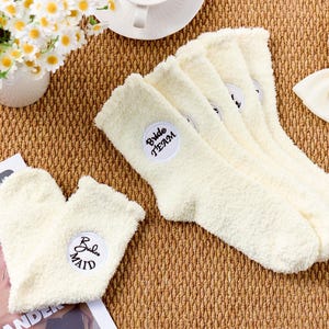 以下が含まれることがあります： クリーム色のふわふわソックス。ハート型のスリッパソックスも含まれています。質感のある表面に表示されています。「Bride Team」と「Bride Maid」の刺繍パッチが付いています。白いリボンとデイジーの花瓶も表示されています。
