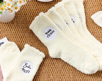 May include: Cream-colored fuzzy socks, including a pair of heart-shaped slipper socks, are displayed on a textured surface. The socks have embroidered patches that read "Bride Team" and "Bride Maid". A white bow and a vase of daisies are also visible.