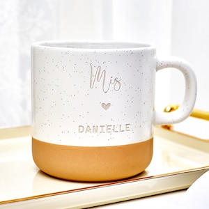 以下が含まれることがあります： タン色のベースと湾曲したハンドルが付いた、白い斑点のあるセラミックマグ。マグには小さなハートの上に「Mis」という文字と、「DANIELLE」という名前が銀色の文字で書かれています。マグは金色の縁取りのトレイに置かれています。