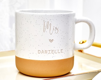 May include: White speckled ceramic mug with a tan base and a curved handle. The mug has the word "Mis" above a small heart and the name "DANIELLE" in silver lettering. The mug is on a gold-trimmed tray.