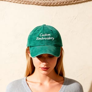 以下が含まれることがあります： 白い「Custom Embroidery」の文字が入った緑色の野球帽。帽子は柔らかい素材でできており、つばがカーブしています。画像に写っている人物は、グレーのVネックセーターを着ています。
