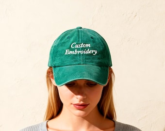 May include: A green baseball cap with the text "Custom Embroidery" in white. The cap is made of a soft material and has a curved brim. The person in the image is wearing a gray v-neck sweater.