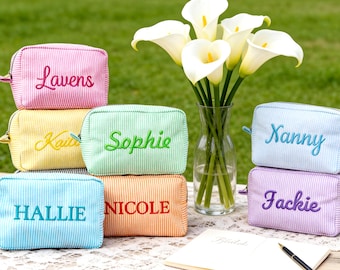 May include: A collection of personalized cosmetic bags in various colors, including pink, yellow, green, blue, orange, and purple. Each bag features a name embroidered in a contrasting color. A bouquet of white calla lilies sits in a glass vase.