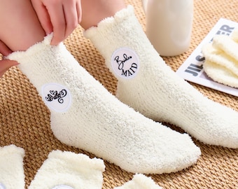 May include: Cream-colored fuzzy socks with ruffled edges. Each sock has a circular patch with embroidered text, including "Bride," "Maid," and "Flower Girl." The socks are displayed on a woven surface, suggesting a cozy, comfortable design.