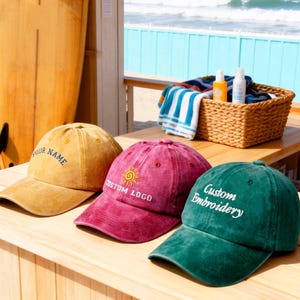 以下が含まれることがあります： マスタードイエロー、バーガンディ、エメラルドグリーンの野球帽が3つ、木製の表面に置かれています。各帽子には「YOUR NAME」、「CUSTOM LOGO」、「Custom Embroidery」というカスタムテキストが入っています。サーフボードとビーチの風景が背景にあります。