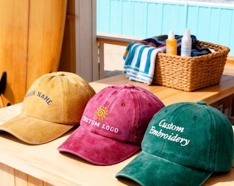May include: Three baseball caps in mustard yellow, burgundy, and emerald green sit on a wooden surface. Each cap has custom text: "YOUR NAME", "CUSTOM LOGO", and "Custom Embroidery". A surfboard and beach scene are in the background.