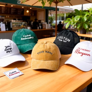 Op de afbeelding: Een verzameling baseballpetten in verschillende kleuren, waaronder wit, groen, zwart en geel. Elke pet heeft aangepaste borduursels met zinnen als "Your Design Here" en "Your Name". De petten worden op een houten tafel getoond.