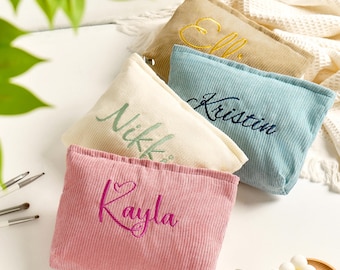 May include: Four corduroy cosmetic bags in beige, light blue, cream, and pink, each embroidered with a name in a contrasting color. Makeup brushes and a wooden tray with a white candle are in the foreground.