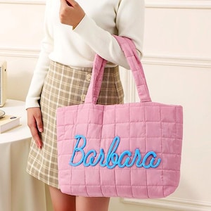 以下が含まれることがあります： ピンク色のキルティングトートバッグ。バッグには青い筆記体で「Barbara」と刺繍されています。バッグは長方形で、格子模様と2つのハンドルが付いています。白いセーターとチェック柄のスカートを着た人がバッグを持っています。
