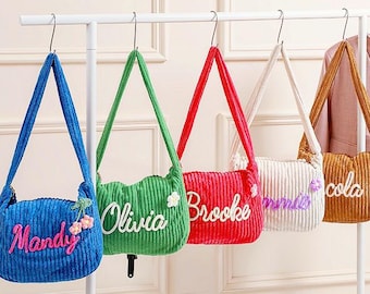 May include: Five corduroy shoulder bags in blue, green, red, white, and tan, each with a different name embroidered on the front. The bags hang from a white rack, with a pink blazer visible in the background.