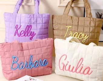 May include: Four quilted tote bags in lavender, tan, pink, and white, each embroidered with a name in a contrasting color. The bags have matching quilted handles and are displayed on a round table with books.