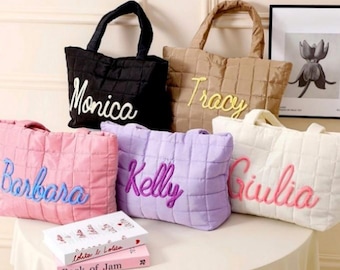 May include: A collection of quilted tote bags in various colors, including black, tan, pink, purple, and white. Each bag features a name embroidered in a contrasting color. The bags are displayed on a round table with books.