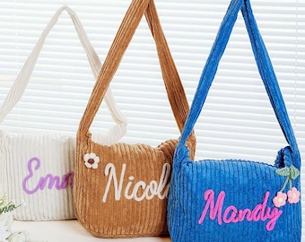 May include: Three corduroy shoulder bags in white, brown, and blue. Each bag has a matching strap and features a name embroidered in a contrasting color: Emma in purple, Nicol in white, and Mandy in pink. The blue bag has a floral embellishment.