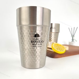 May include: A stainless steel tumbler with a textured surface and the text "ROMEO BEST MAN 10-10-2025" printed on the front. The tumbler has a hammered finish and a straight-sided design. A second tumbler is visible in the background.