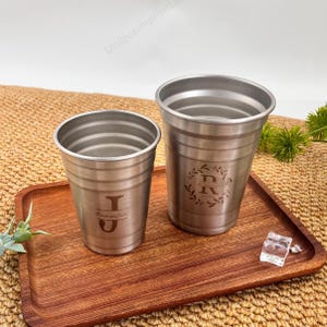 Może przedstawiać: Dwa kubki ze stali nierdzewnej z wygrawerowanymi inicjałami i tekstem. Jeden kubek ma literę "J" i tekst "Roses and Juleps". Drugi kubek ma literę "R" wewnątrz wzoru wieńca laurowego. Oba kubki znajdują się na drewnianej tacy.