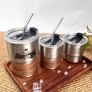 Peut inclure: Trois gobelets en acier inoxydable avec couvercles transparents et pailles en métal sont présentés sur un plateau en bois. Chaque gobelet a une base en bois. L'un présente un motif de montagne et de camping-car, un autre un voilier, et le troisième porte le nom "John".