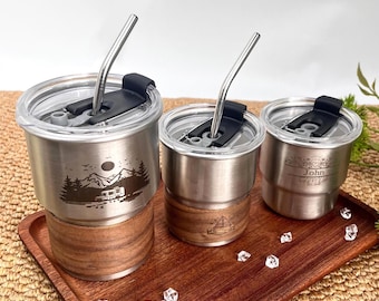 May include: Three stainless steel tumblers with clear lids and metal straws sit on a wooden tray. Each tumbler has a wooden base. One tumbler features a mountain and camper design, another a sailboat, and the third has the name "John".