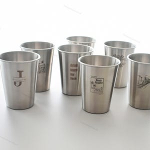 Może przedstawiać: Kolekcja kubków do piwa ze stali nierdzewnej z różnymi wzorami. Jeden kubek ma literę "J" z tekstem, inny mówi "drink more mr jack", a inny ma scenę plażową. Kubki są cylindryczne i mają srebrne wykończenie.