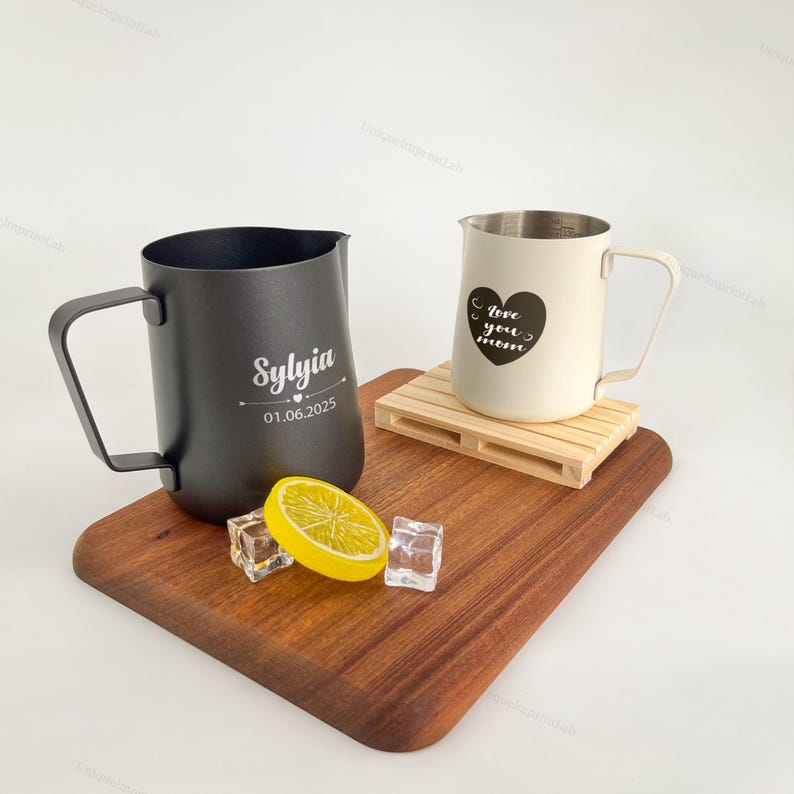 May include: Two metal pitchers, one black with "Sylvia" and a date, the other cream-colored with a heart design and "love you mom" text. They sit on a wooden board with a lemon slice and ice cubes. The cream pitcher rests on a small wooden pallet.