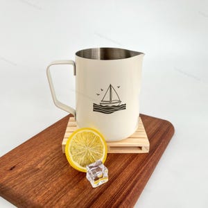 May include: Cream-colored metal pitcher with a sailboat graphic. The pitcher has a silver rim and a handle. It sits on a small wooden pallet, next to a lemon slice and an ice cube on a wooden cutting board.