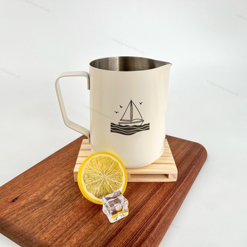 May include: Cream-colored metal pitcher with a sailboat graphic. The pitcher has a silver rim and a handle. It sits on a small wooden pallet, next to a lemon slice and an ice cube on a wooden cutting board.