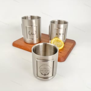 Puede incluir: Tres tazas de acero inoxidable con diseños grabados. Una taza presenta la letra "A" dentro de una corona de laurel, otra tiene "SMS", y la tercera dice "Welcome to our home, the Romeo and Juliet family". Una rodaja de limón descansa sobre una tabla de madera.