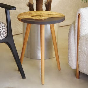 Puede incluir: Una pequeña mesa auxiliar redonda con una tapa de madera natural y un borde gris oscuro. La mesa tiene tres patas de madera de color claro. Una silla negra con un cojín estampado y un sillón blanco están en el fondo. Una palmera en maceta está detrás de la mesa.
