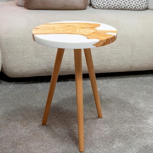 Könnte beinhalten: Kleiner, runder Beistelltisch mit einer weißen Tischplatte, die mit natürlicher Holzmaserung eingelegt ist. Der Tisch hat drei Holzbeine. Der Tisch hat einen Durchmesser von ca. 46 cm und ist 51 cm hoch. Der Tisch steht vor einem hellen Sofa.