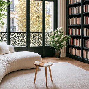 May include: A cozy living room with a curved white sofa, a small round wooden side table, and a large bookshelf filled with books. A large window with decorative ironwork overlooks a tree-lined street.