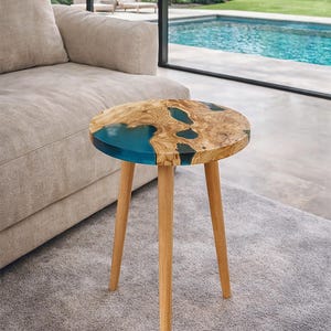 Może przedstawiać: Mały, okrągły stolik boczny z drewnianym blatem i trzema drewnianymi nogami. Blat ma naturalny wzór słojów drewna z sekcją z przezroczystej niebieskiej żywicy. Stół ma średnicę około 50 cm.