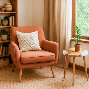 May include: An orange armchair with a cream-colored macrame pillow sits next to a small wooden side table with a plant. A wooden bookshelf filled with books and plants is in the background. The room has a warm, inviting atmosphere.