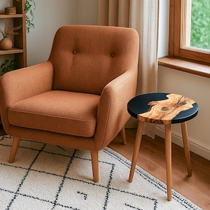 May include: An orange-brown armchair with a buttoned back sits next to a small side table. The table has a black top with a natural wood grain design and three wooden legs. A wooden bookshelf and a patterned rug are also visible.