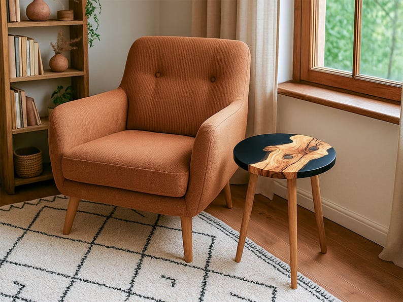 May include: An orange-brown armchair with a buttoned back sits next to a small side table. The table has a black top with a natural wood grain design and three wooden legs. A wooden bookshelf and a patterned rug are also visible.