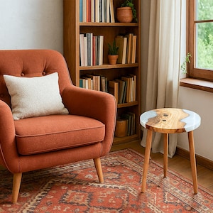 May include: An orange armchair with a white pillow sits next to a small wooden side table with a light-colored epoxy resin top. A wooden bookshelf filled with books and plants is in the background. A patterned rug covers the floor.