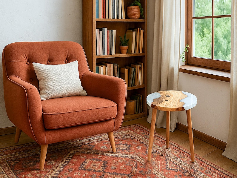 May include: An orange armchair with a white pillow sits next to a small wooden side table with a light-colored epoxy resin top. A wooden bookshelf filled with books and plants is in the background. A patterned rug covers the floor.