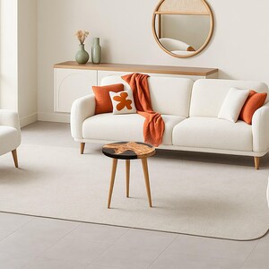 May include: A modern living room setting featuring a white sofa and armchair with orange and white accent pillows and a matching throw blanket. A round wooden coffee table with a black top and three legs sits in front of the sofa. A round mirror hangs on the wall.