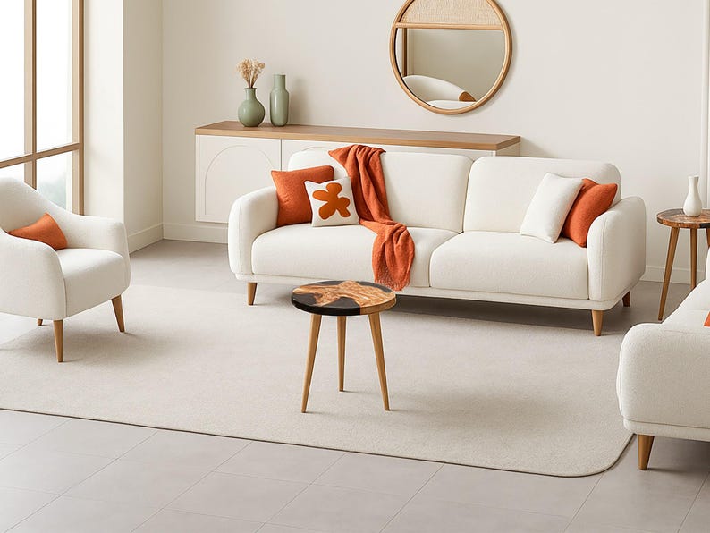 May include: A modern living room setting featuring a white sofa and armchair with orange and white accent pillows and a matching throw blanket. A round wooden coffee table with a black top and three legs sits in front of the sofa. A round mirror hangs on the wall.