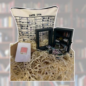 May include: A white pillow with a black and white calendar design that says "Date Due". The pillow is surrounded by shredded paper and a small black box with a glass front filled with miniature books. There is also a small pink book on a white card.
