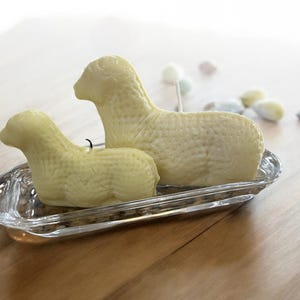 May include: Two white ceramic lamb shaped candles in a clear glass dish. The candles are in a sitting position and have a textured surface.