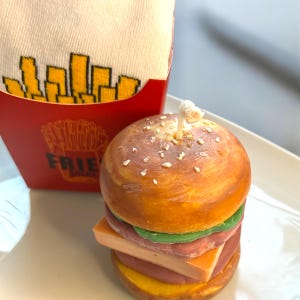 May include: A yellow and brown hamburger candle with a white wick. The candle is on a white plate. A red and white box with the word "FRIES" is in the background.