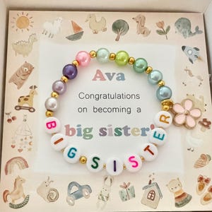 Może przedstawiać: Biała kartka z kolorowym koralikowym bransoletką i tekstem "Ava Gratulacje z okazji zostania starszą siostrą". Kartka jest ozdobiona fantazyjną ilustracją zwierząt i przedmiot&oacute;w.